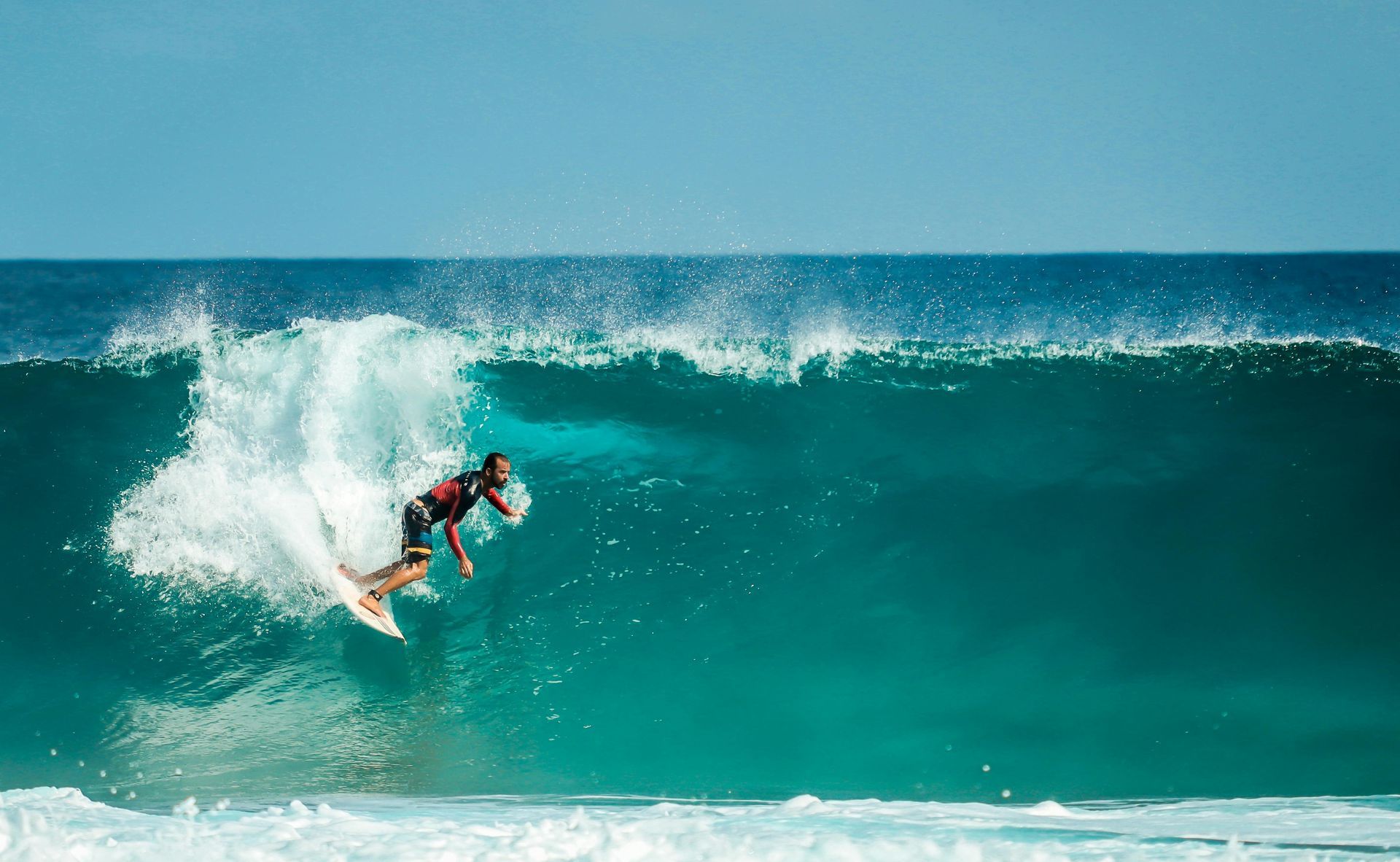 Surfer carves a wave, spray erupting. Turquoise water, blue sky.