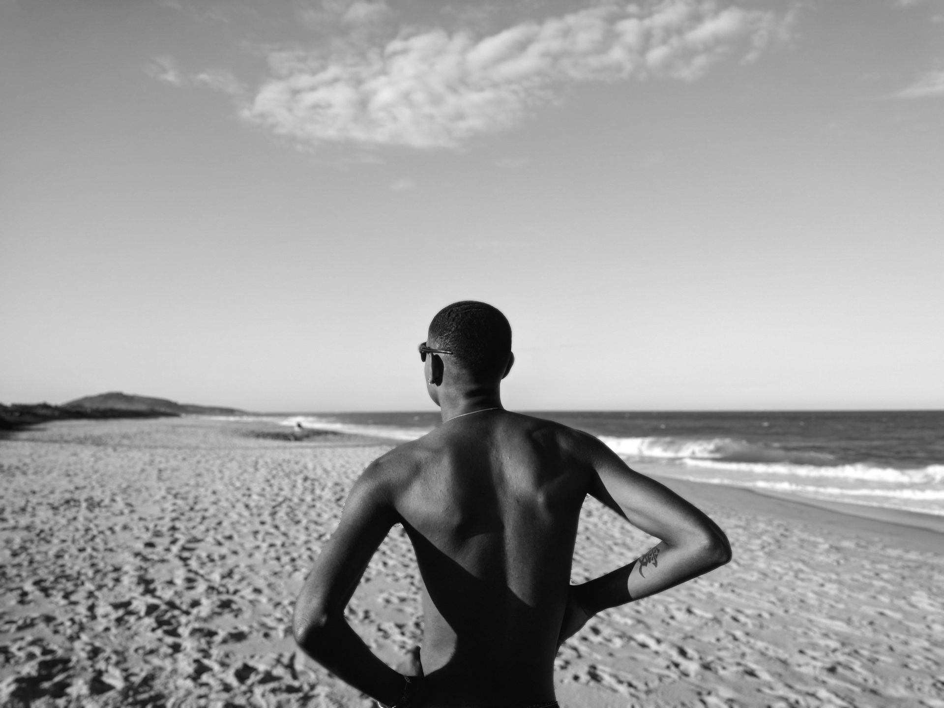 A person stands on a sandy beach, arms on hips, facing the ocean under a partly cloudy sky.