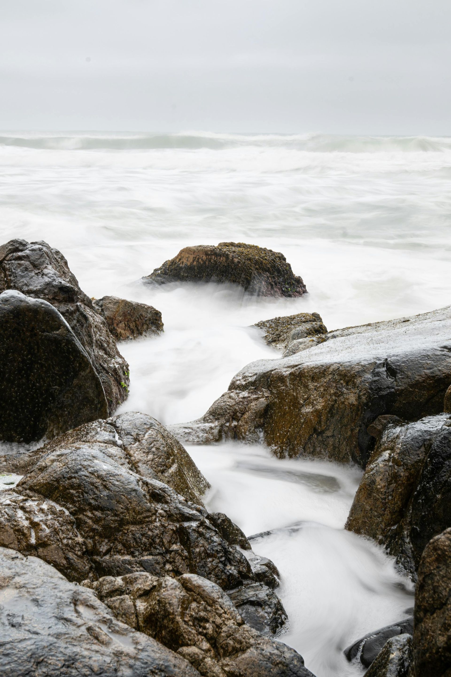Waves crashing over dark, wet rocks on a cloudy day.