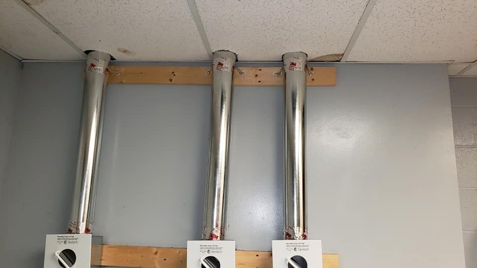 Three silver pipes run from the ceiling to white electrical boxes on a gray wall, attached to a wooden board.
