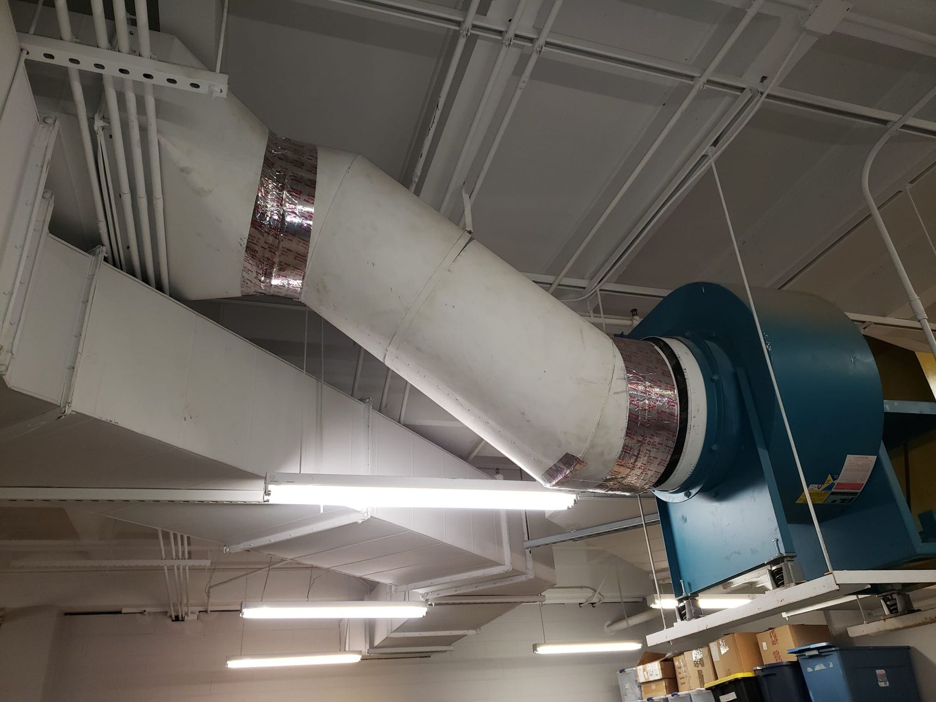 Ceiling-mounted industrial ventilation system with rectangular and cylindrical ducts, wrapped in silver tape. Blue fan on the right.