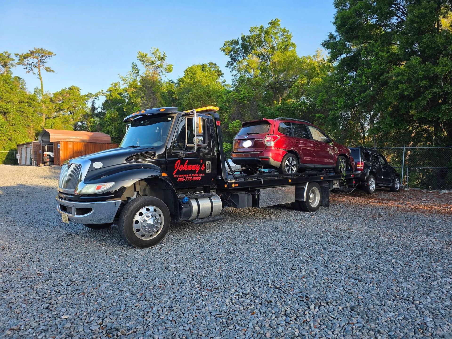 Tow truck with two cars, one red, one black, parked on gravel, sunny outdoor setting.
