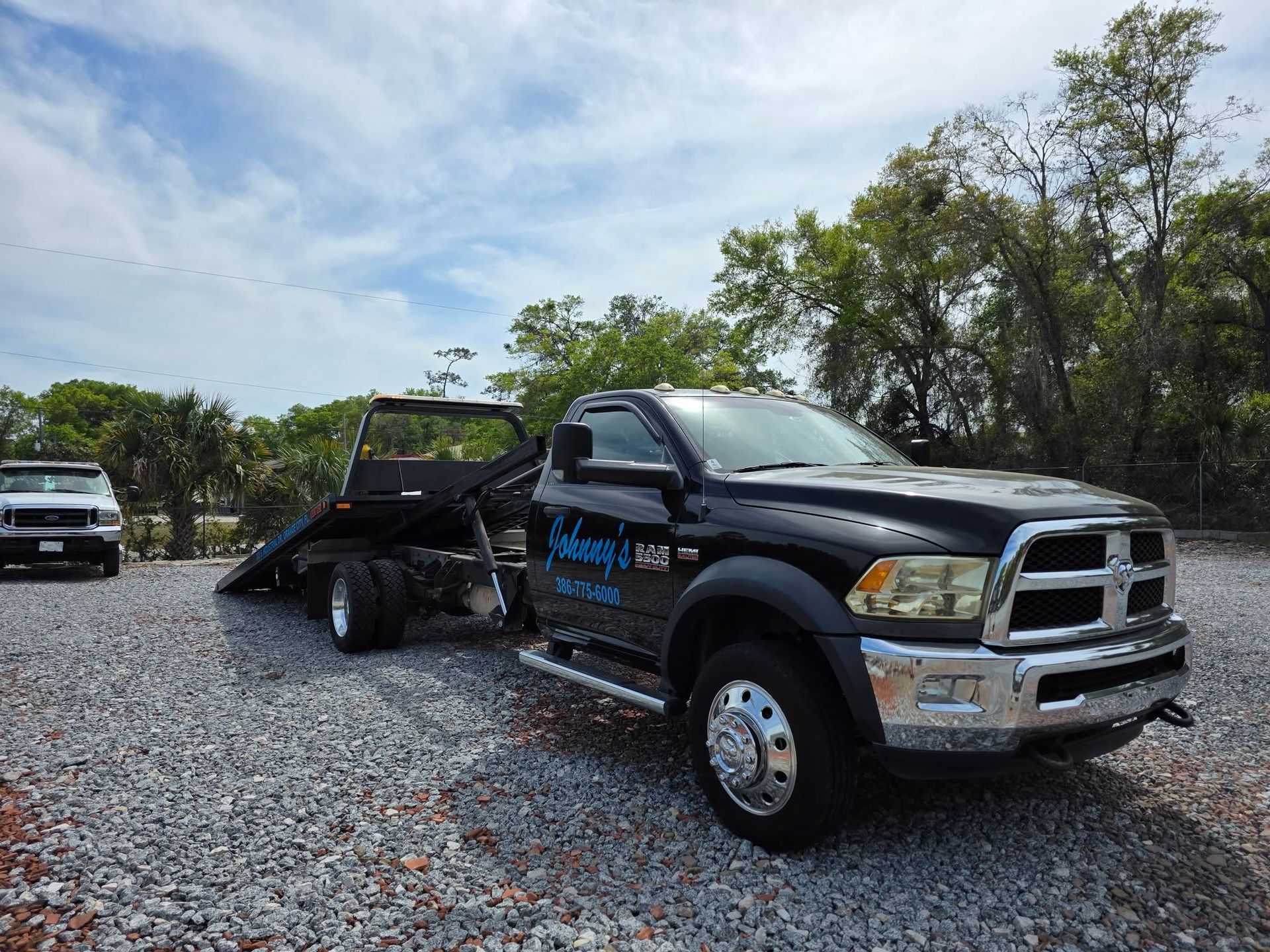 Black tow truck with flatbed in a gravel lot under a blue sky, another truck visible in the background.