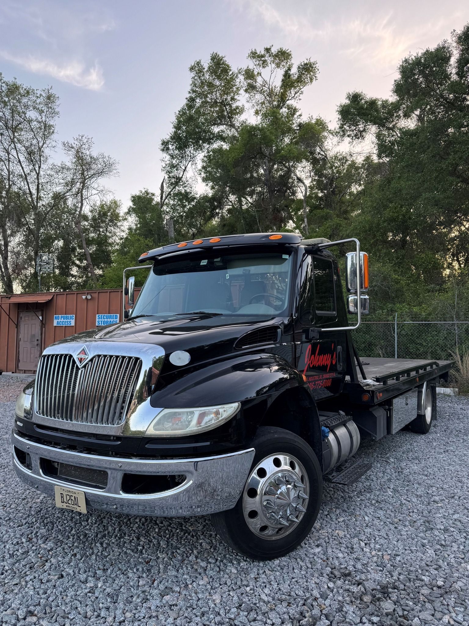 Black tow truck parked on gravel, facing left, with a chrome grill and wheels, against a backdrop of trees.