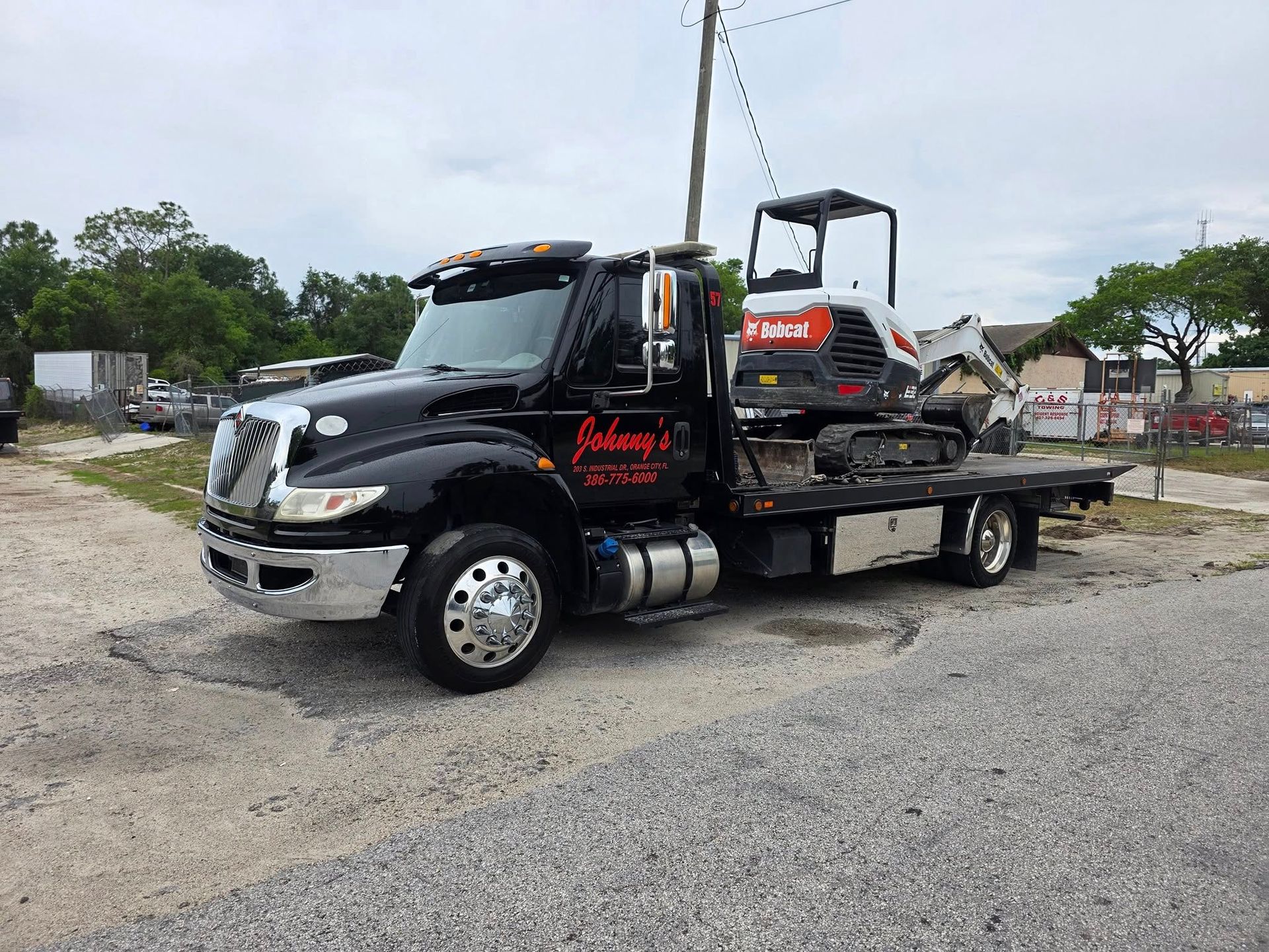 Black tow truck with Bobcat excavator on flatbed, parked on gravel.