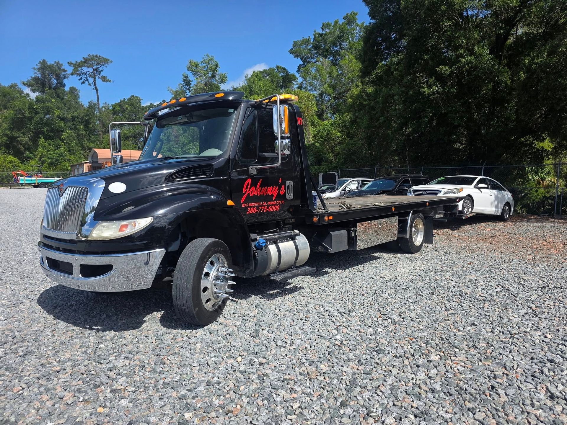 Black tow truck with a white car on a flatbed, parked on gravel under a blue sky.