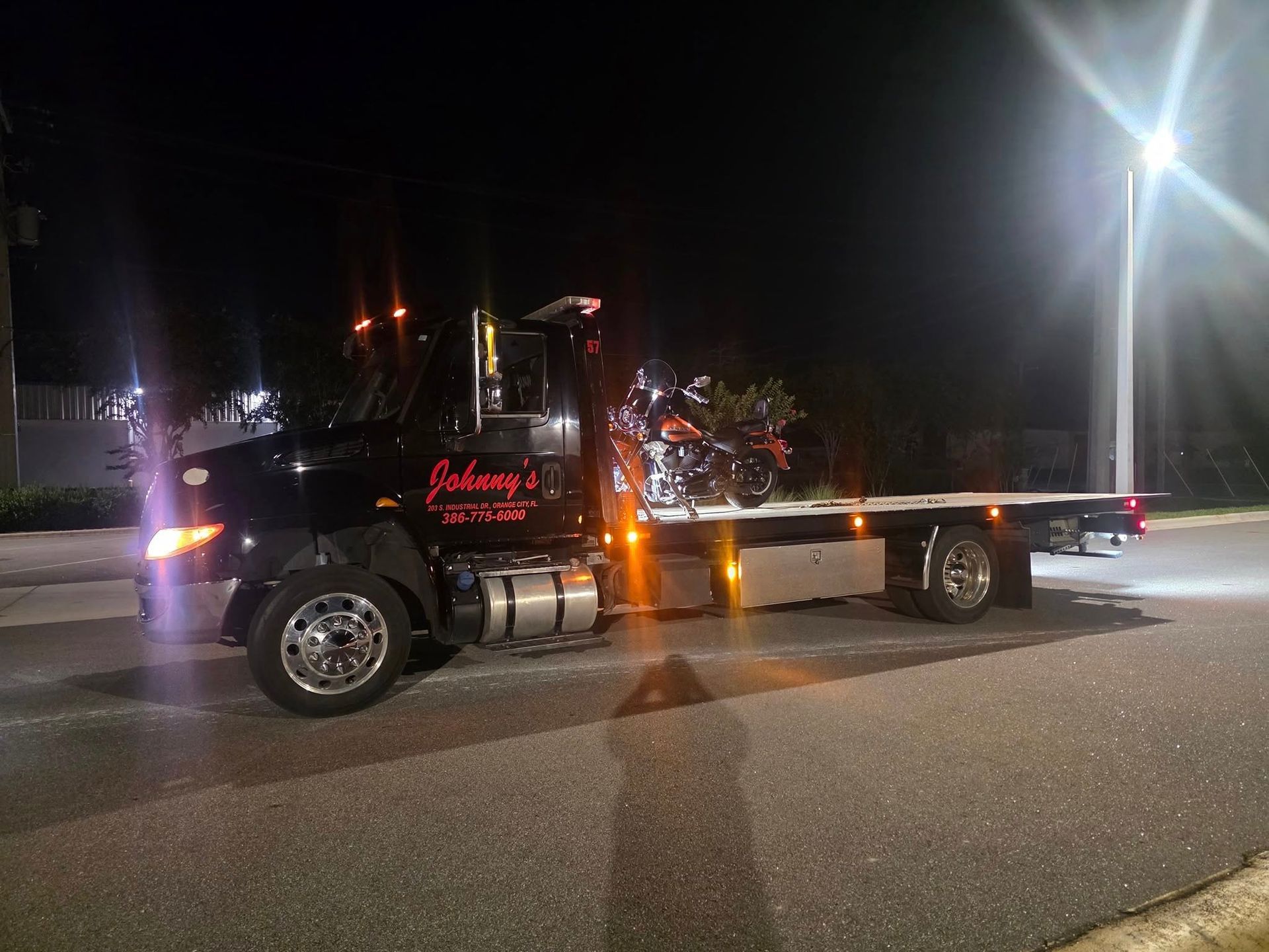 Black tow truck at night, towing a motorcycle. The truck is lit by streetlights.