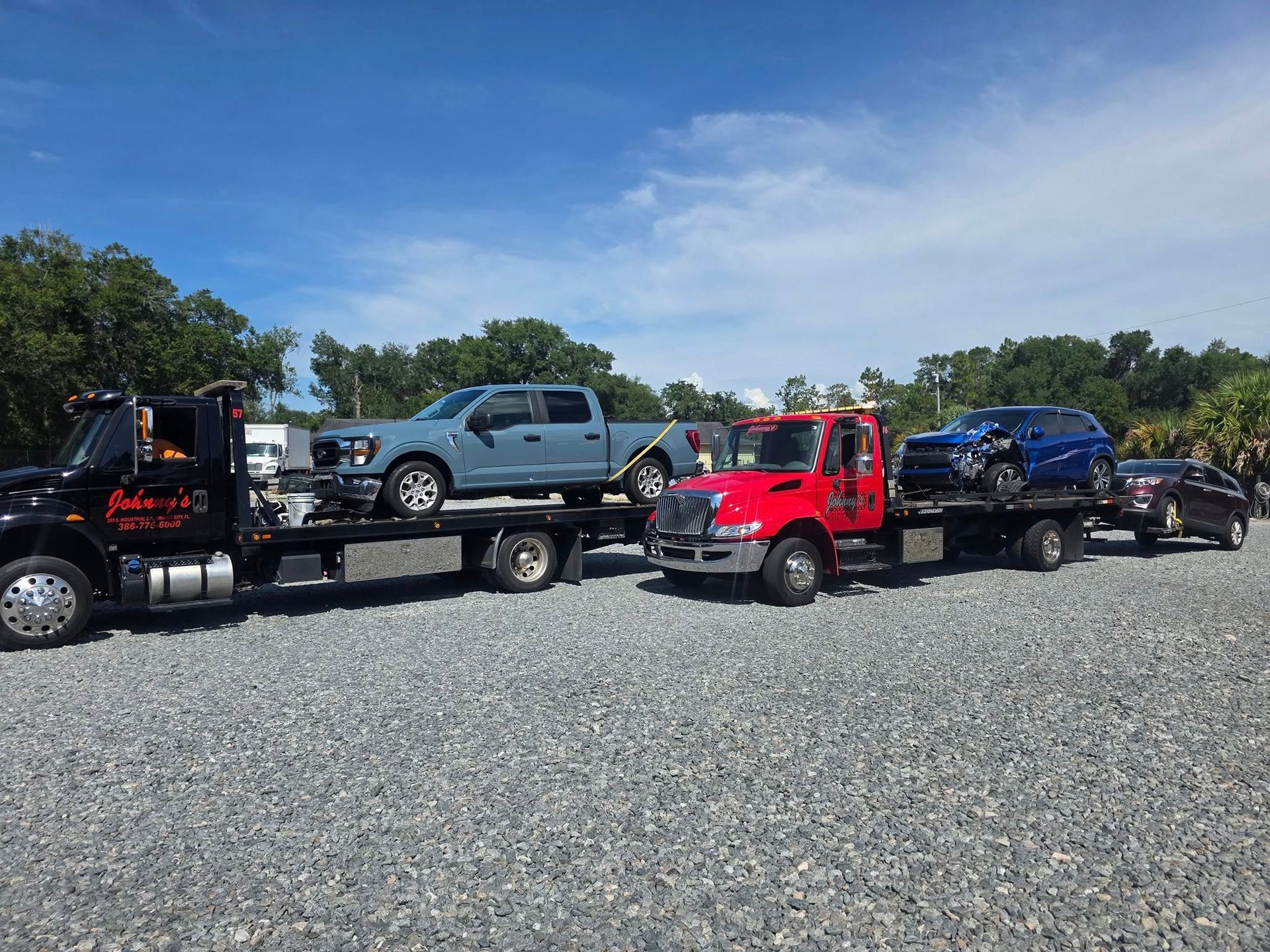 Three tow trucks carrying vehicles on a gravel lot under a blue sky.