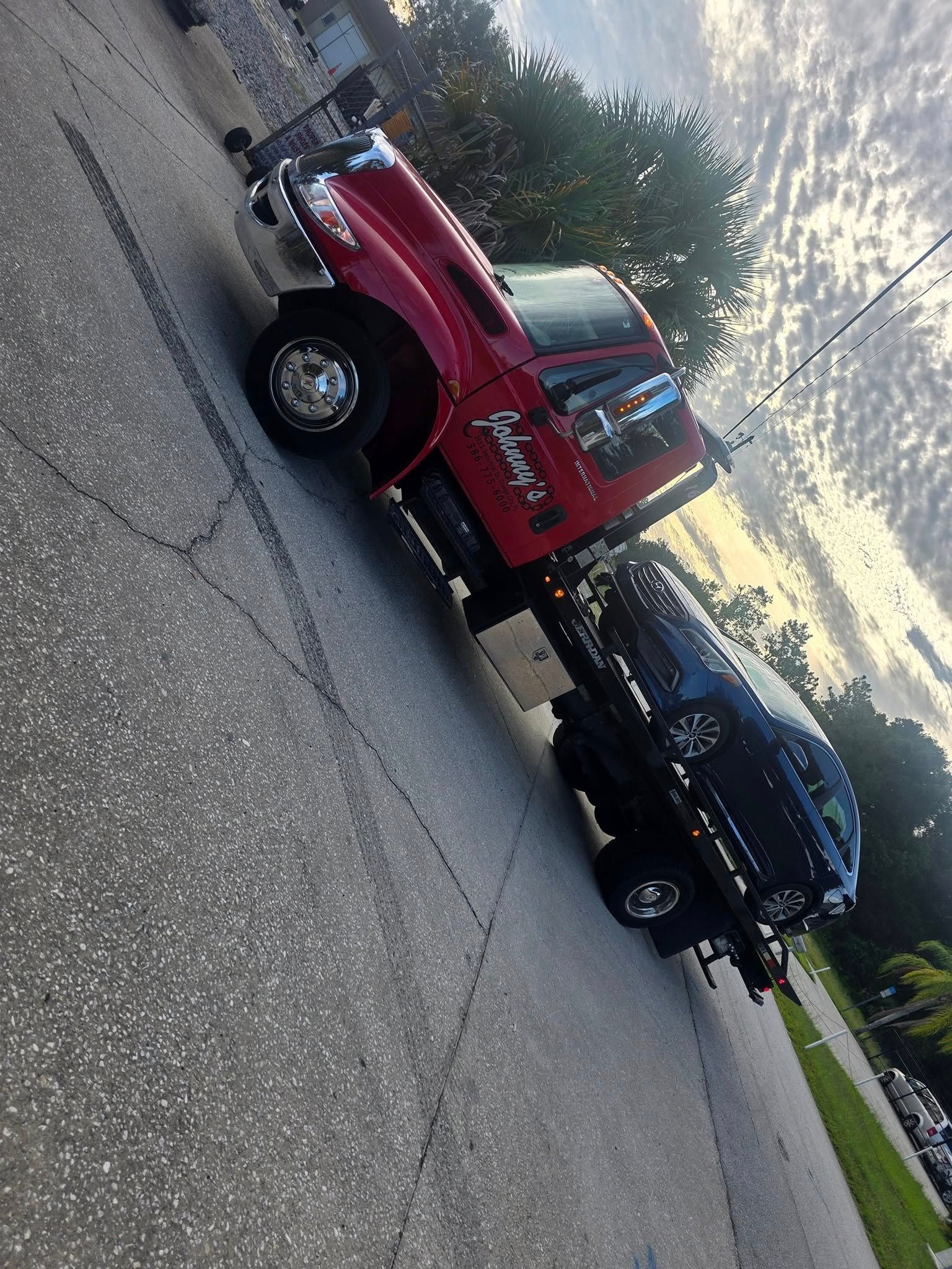 Red tow truck hauling a black car on asphalt driveway; cloudy sky.