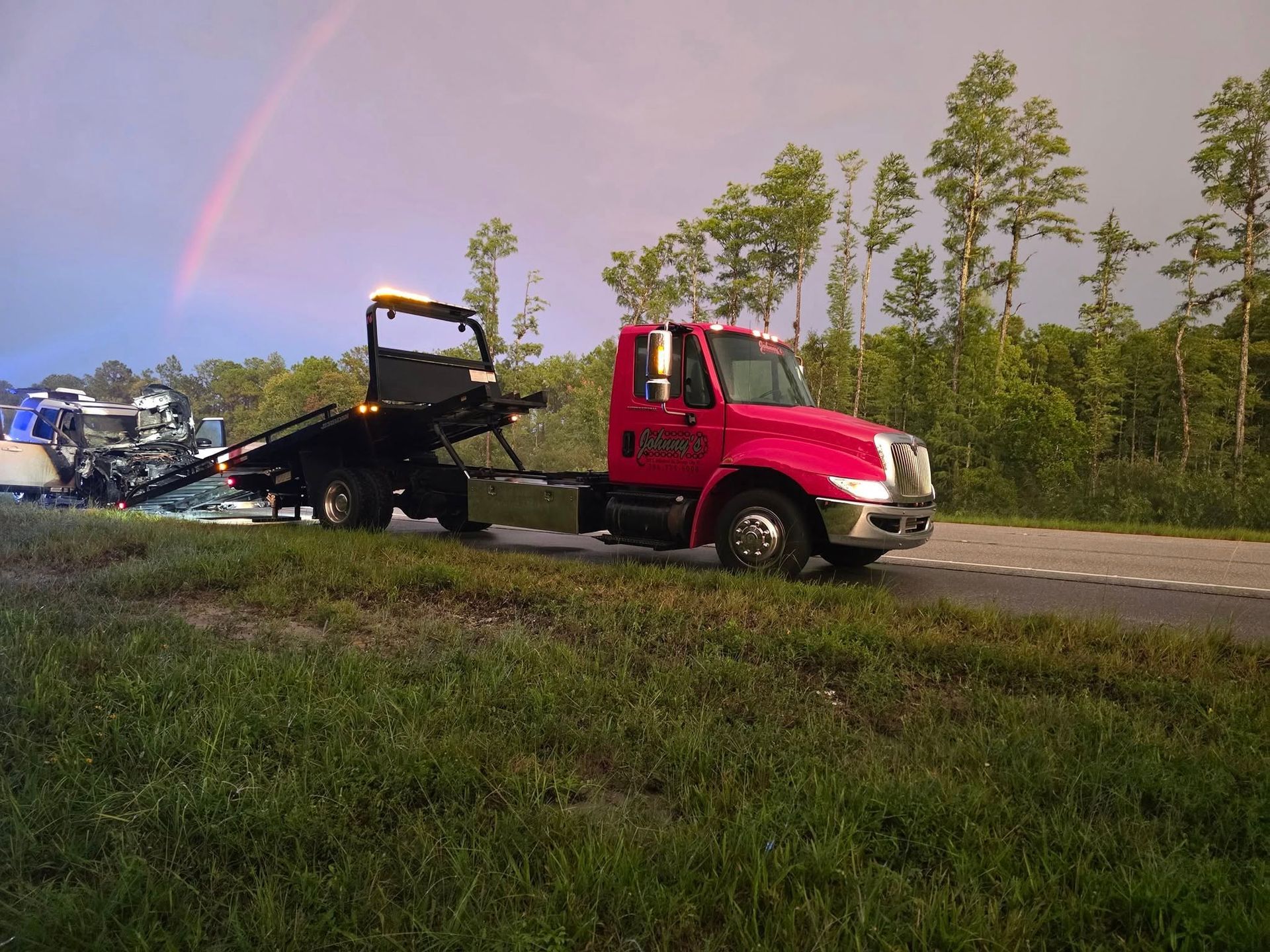 A tow truck in the rain, hauling a crashed car on a highway with a rainbow in the sky.