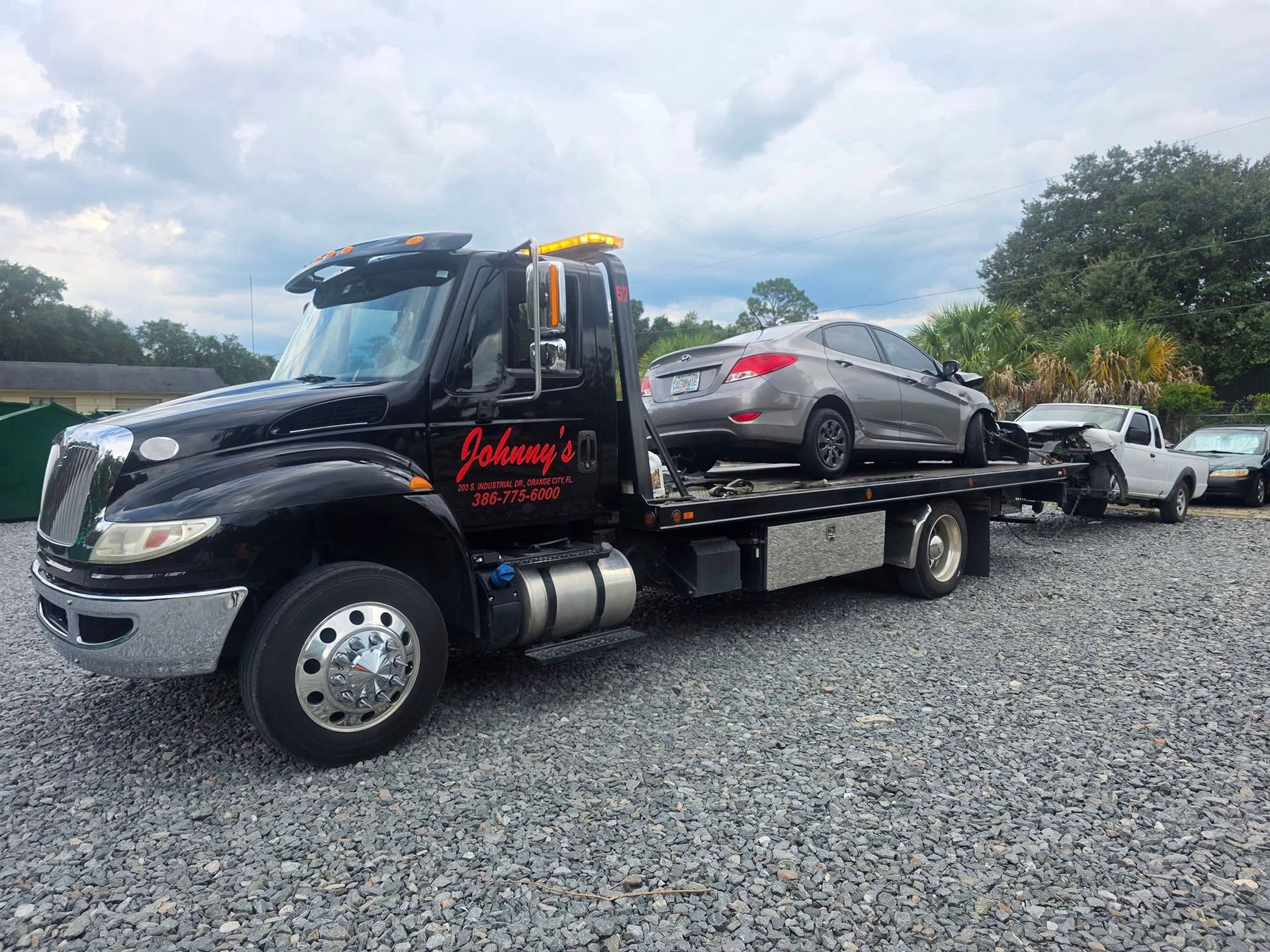Black tow truck transporting a silver car on a gravel lot; blue sky background.