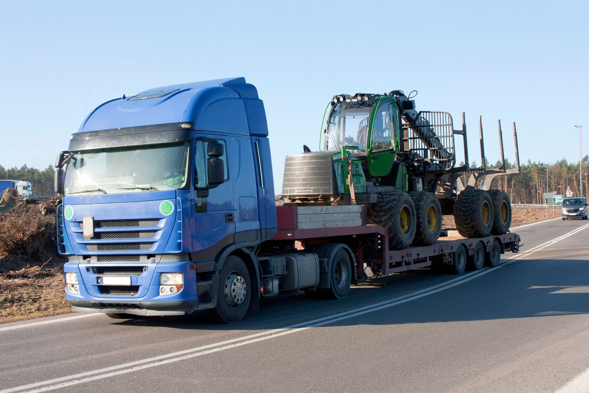 Blue semi-truck hauling a green tractor on a flatbed trailer, driving on a sunny road.