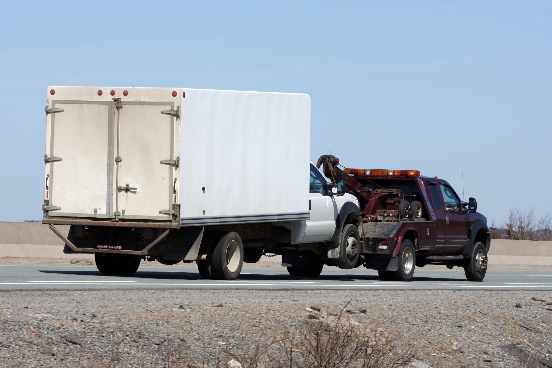 A white truck is being towed by a maroon tow truck on a highway under a blue sky.
