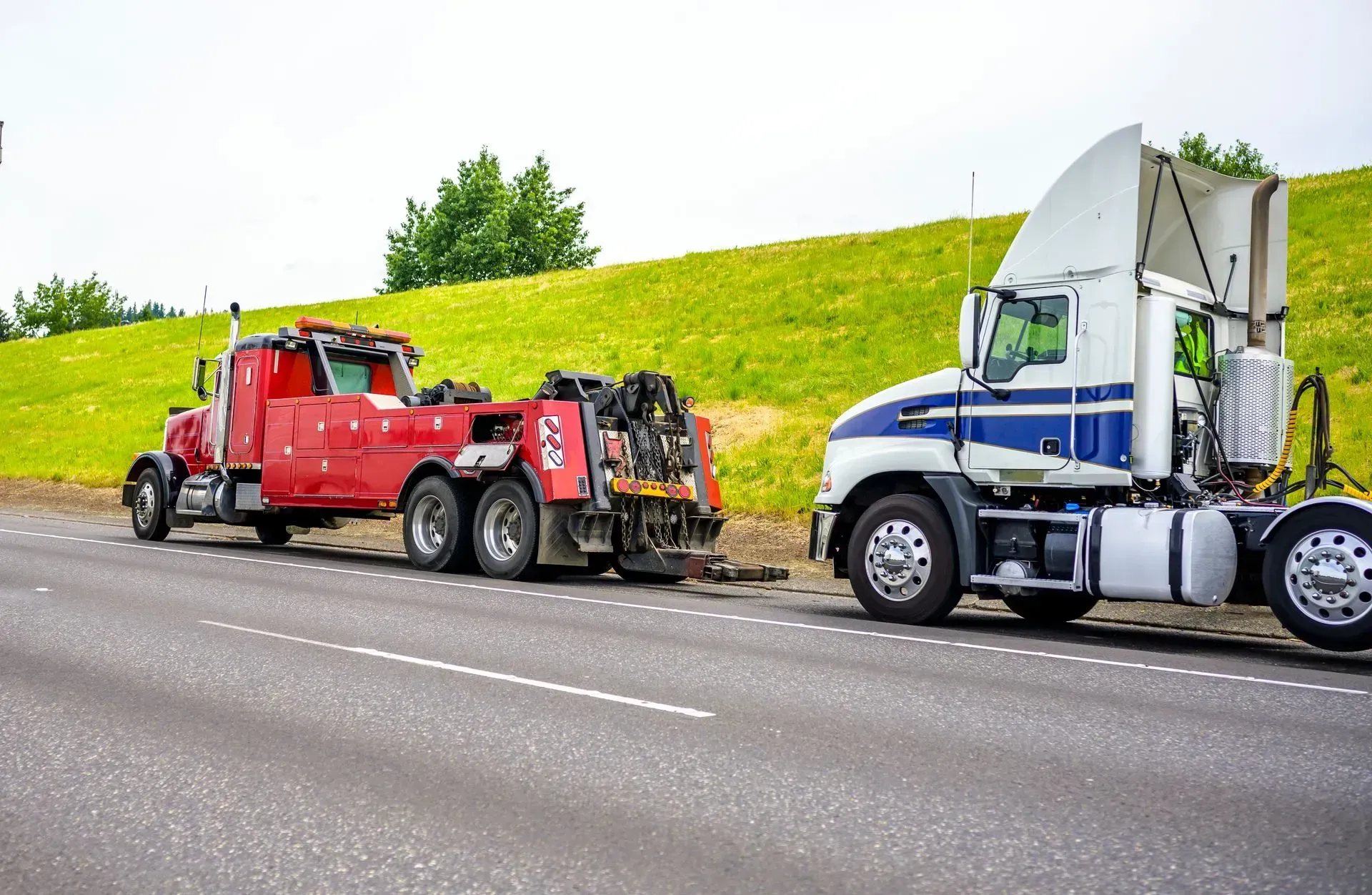 Red tow truck towing a white and blue semi-truck on the side of a road, with a green grassy hill in the background.