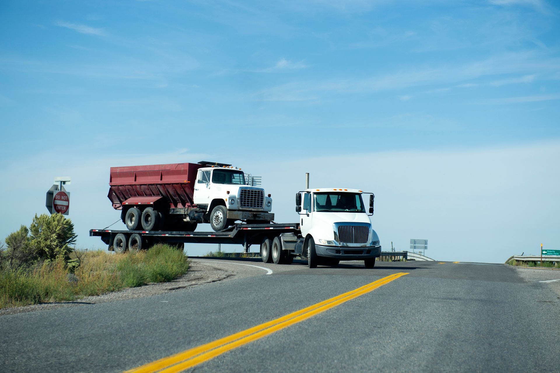 White semi-truck transporting a red tanker truck on a flatbed trailer on a sunny road.