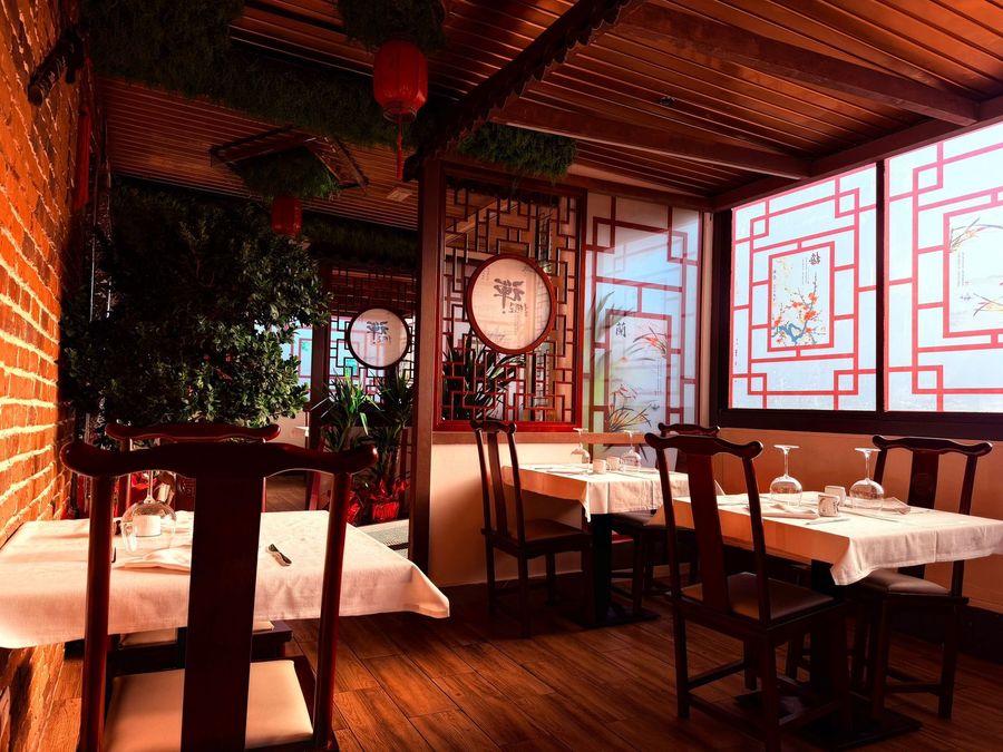 Accogliente interno di un ristorante con sedie in legno, tovaglie bianche, fiori rossi e pannelli decorativi in ​​stile cinese.
