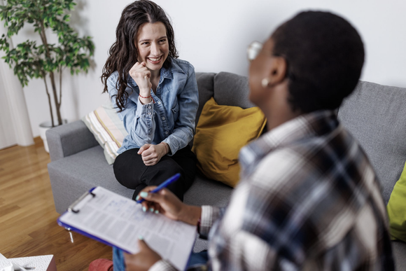 A professional in a plaid shirt takes notes on a clipboard while seated across from a smiling person on a gray couch.