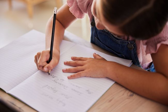 A person writing in a notebook with a black pencil while sitting at a wooden desk.
