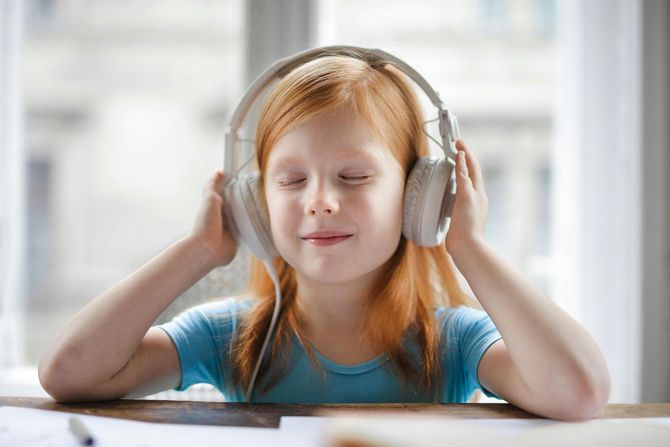 A child with light red hair wearing large white over-ear headphones, eyes closed, resting their hands on the ear cups.