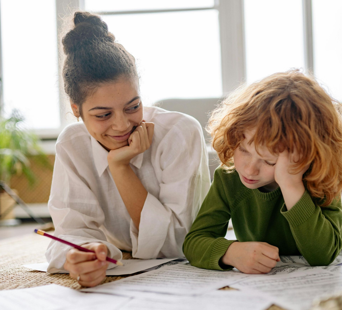 A smiling person holding a pencil watches a person in a green shirt working on a document on the floor.