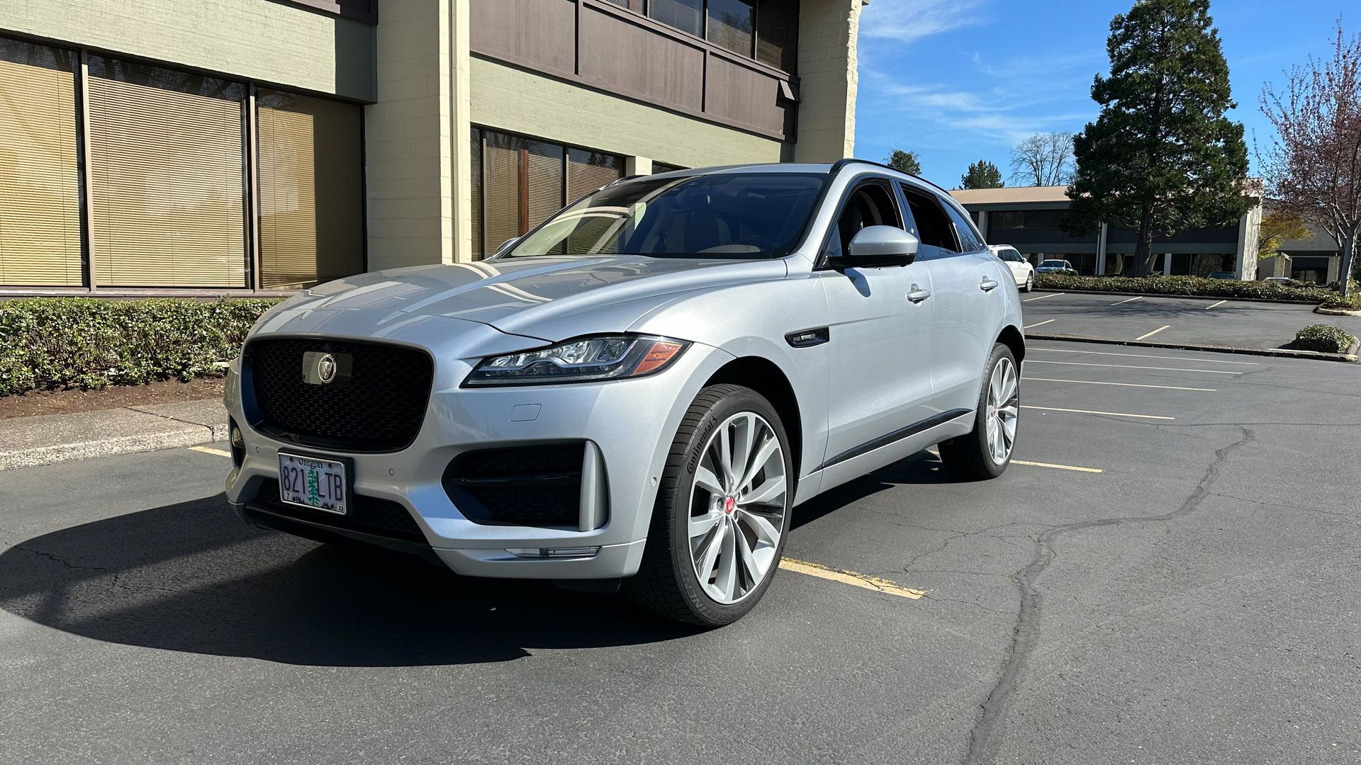 A silver jaguar f pace is parked in a parking lot in front of a building.