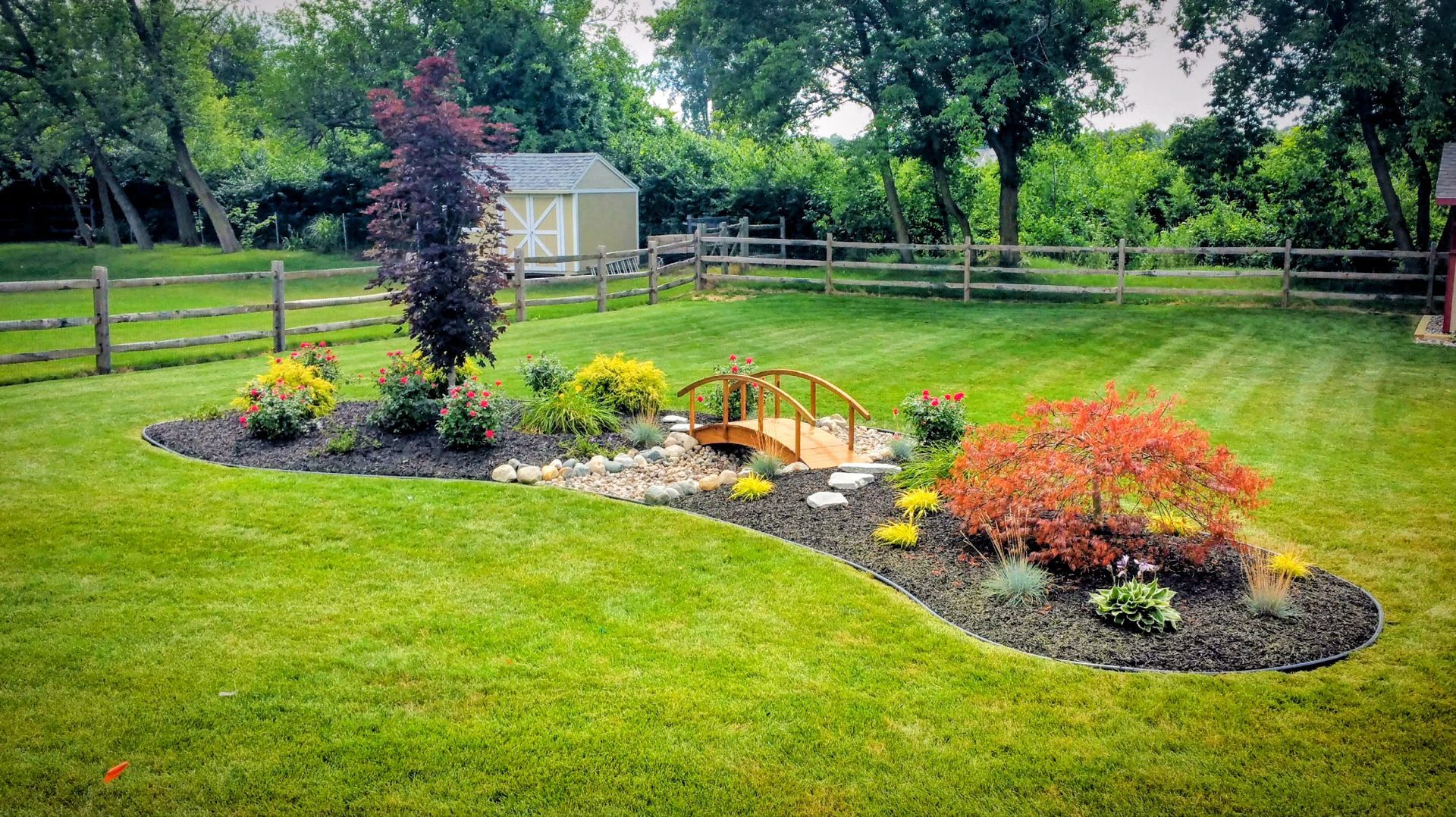 A curved garden bed with a small wooden bridge, colorful shrubs, and mulch in a green, mowed backyard.