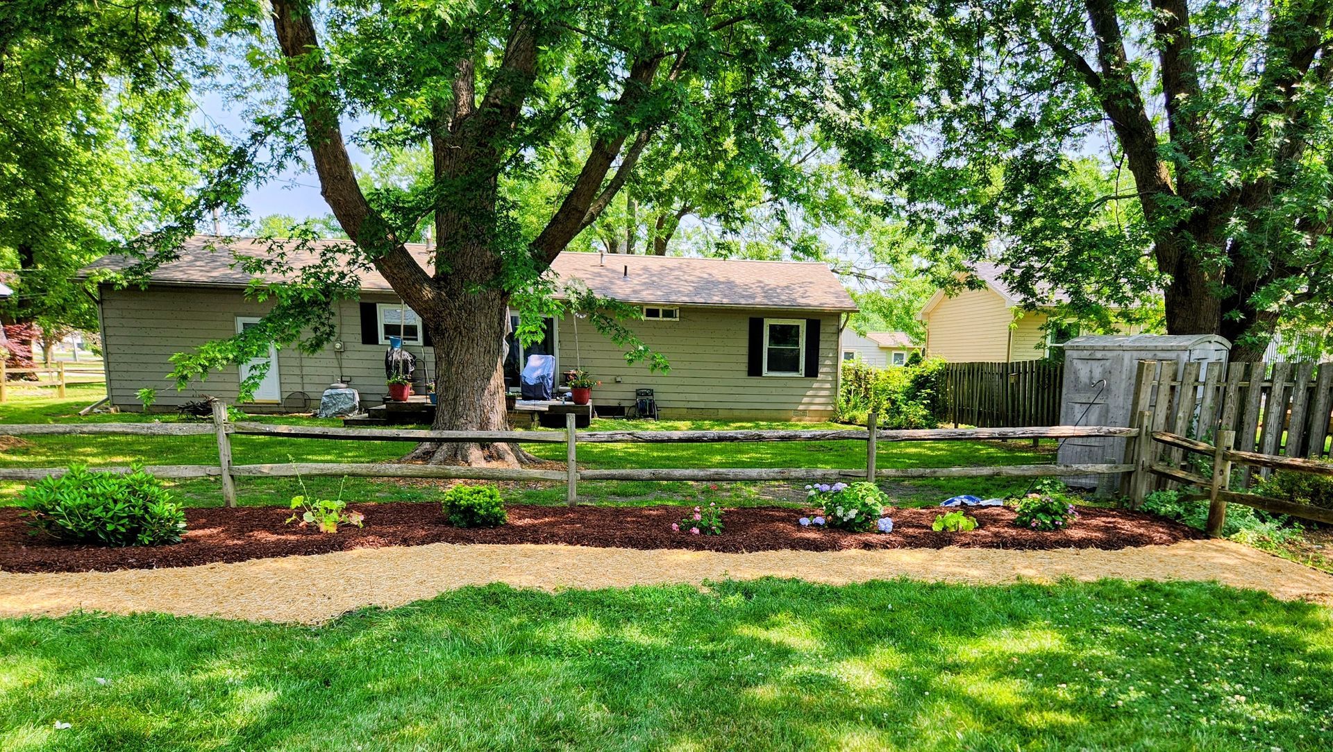A landscaped flower bed with mulch and small plants sits in front of a wooden fence, large trees, and a beige house.