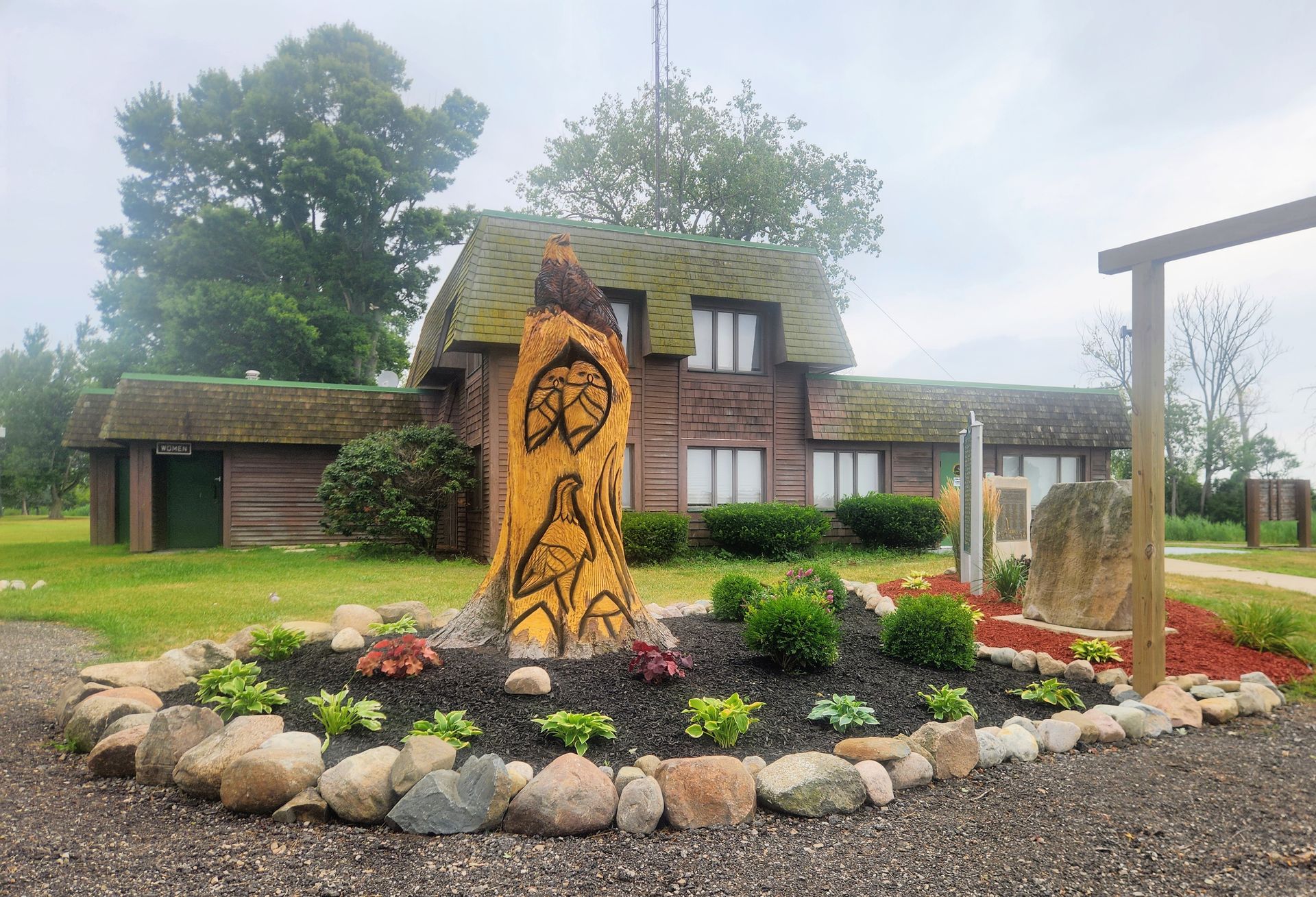 A carved wooden totem stands in a mulched garden bed in front of a quaint cottage with a green roof.