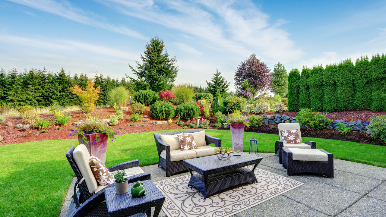 A patio seating area with a sofa, two chairs, and a coffee table on a patterned rug in a lush, landscaped backyard.