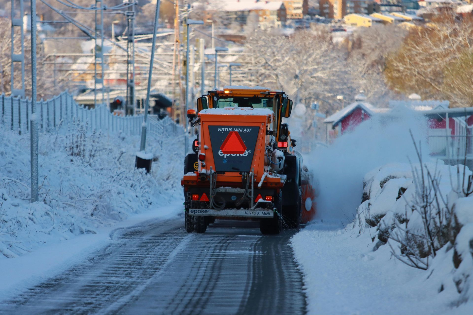 An orange snowblower truck clears snow from a narrow, icy road in a residential area on a bright winter day.