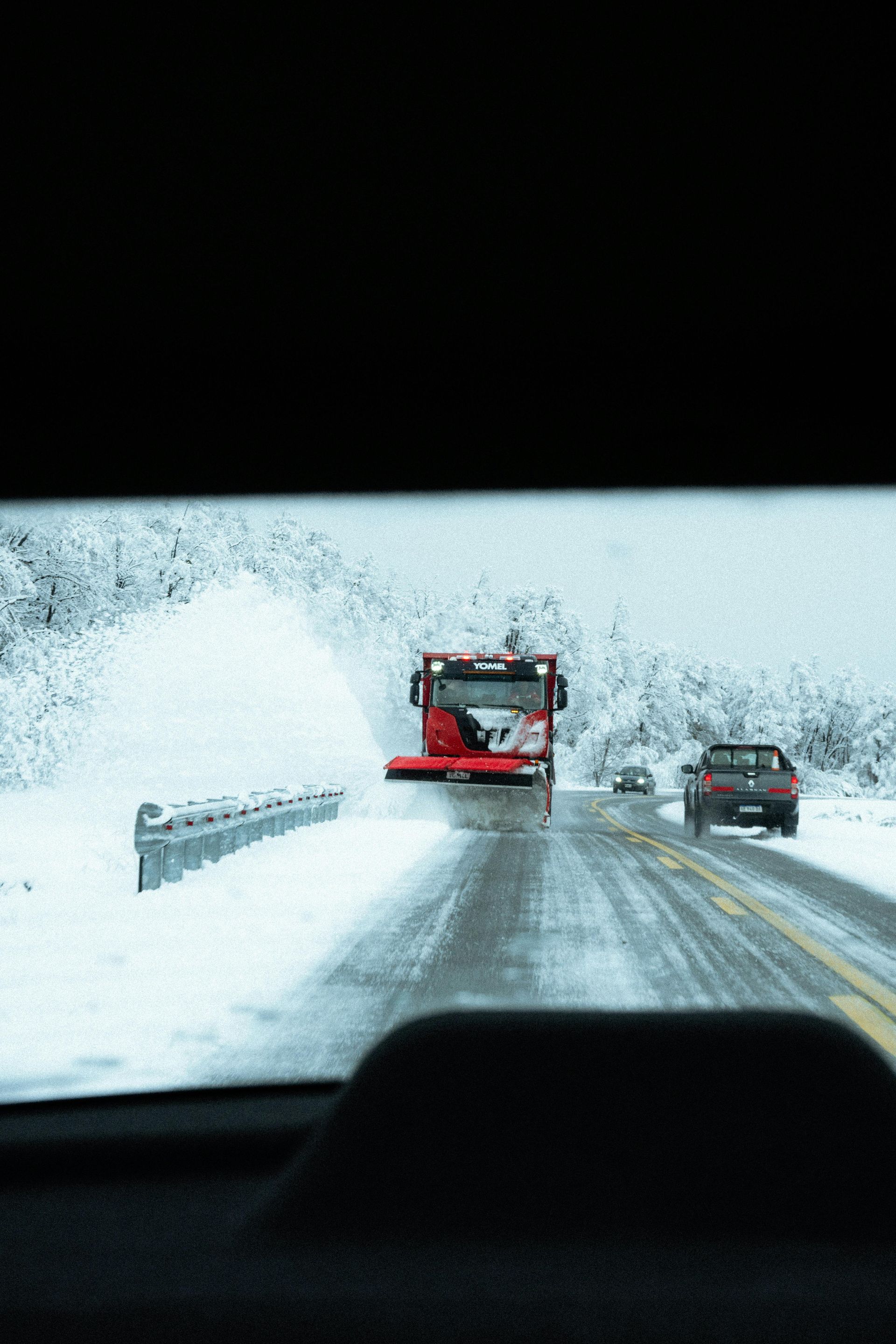 A red snowplow clears a snow-covered road, seen from the perspective of a driver following behind.