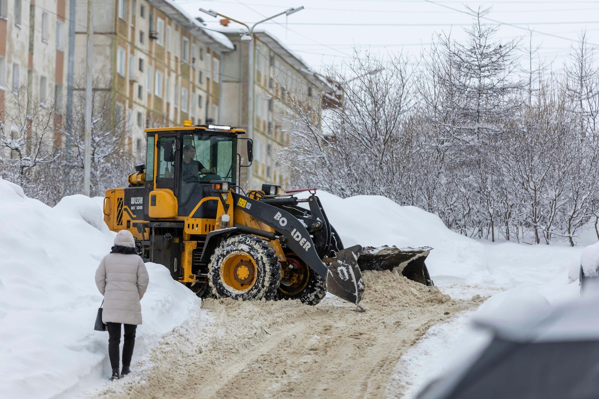 A yellow front-end loader clears heavy snow from a street near an apartment building as a person walks nearby.