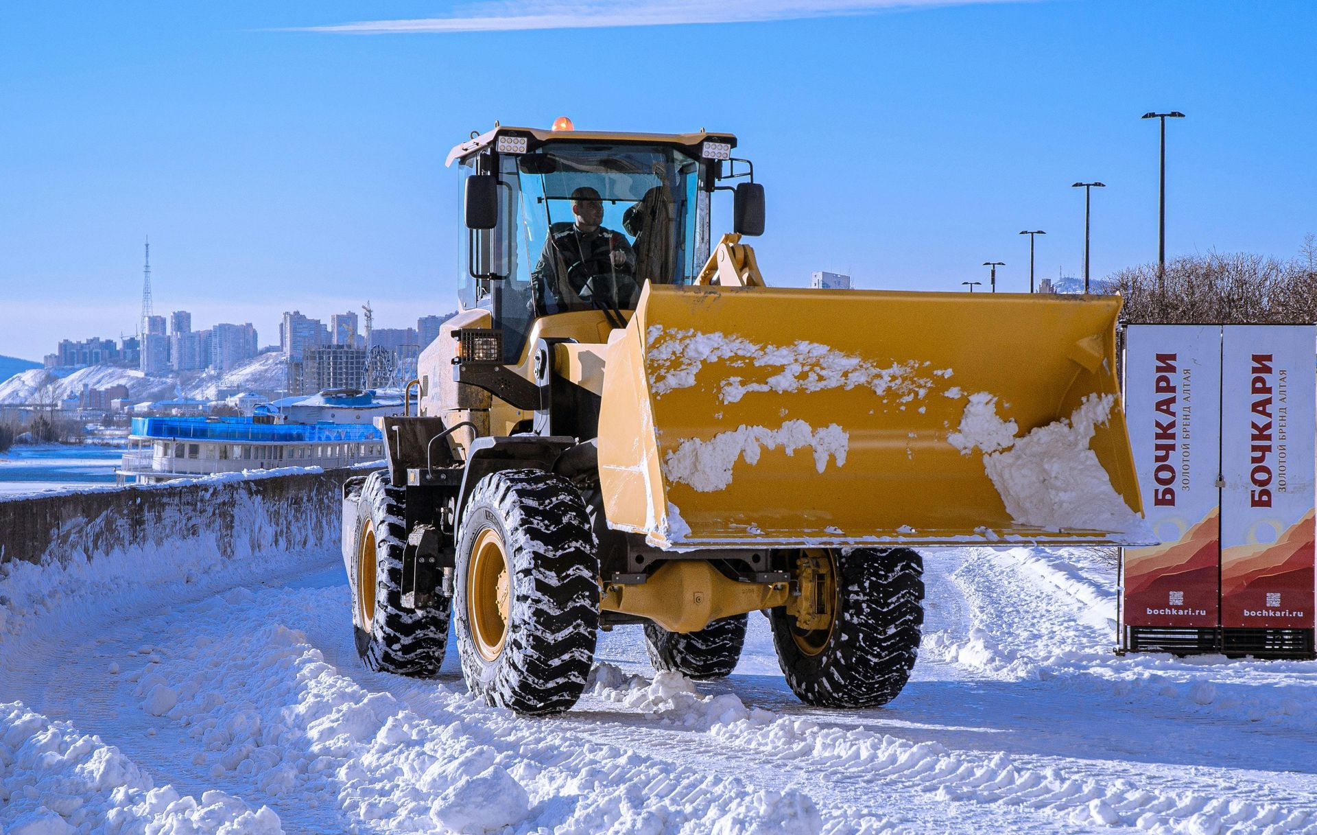 A yellow front-end loader moves snow on a sunny winter day near a waterfront with city buildings in the background.