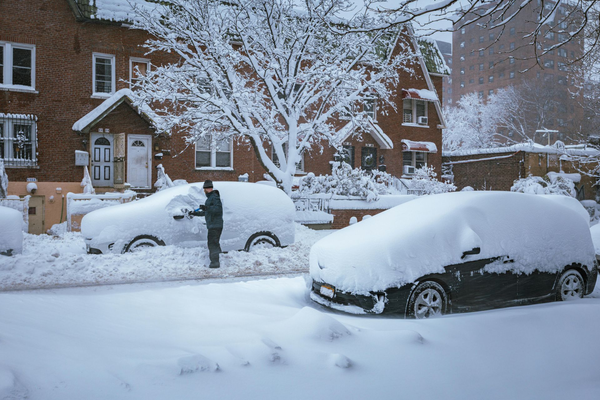 A person clears deep snow from a car parked on a residential street lined with snow-covered brick houses and trees.