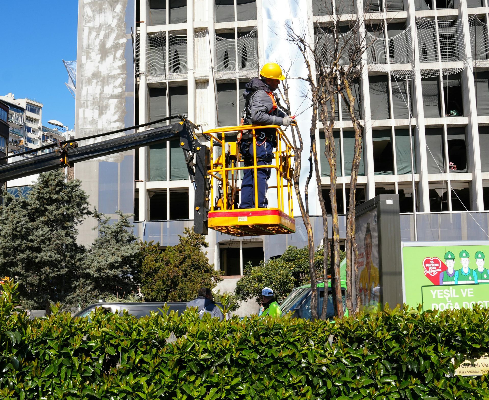 A worker in a yellow hard hat trims a tree from an elevated work platform in front of a modern glass office building.