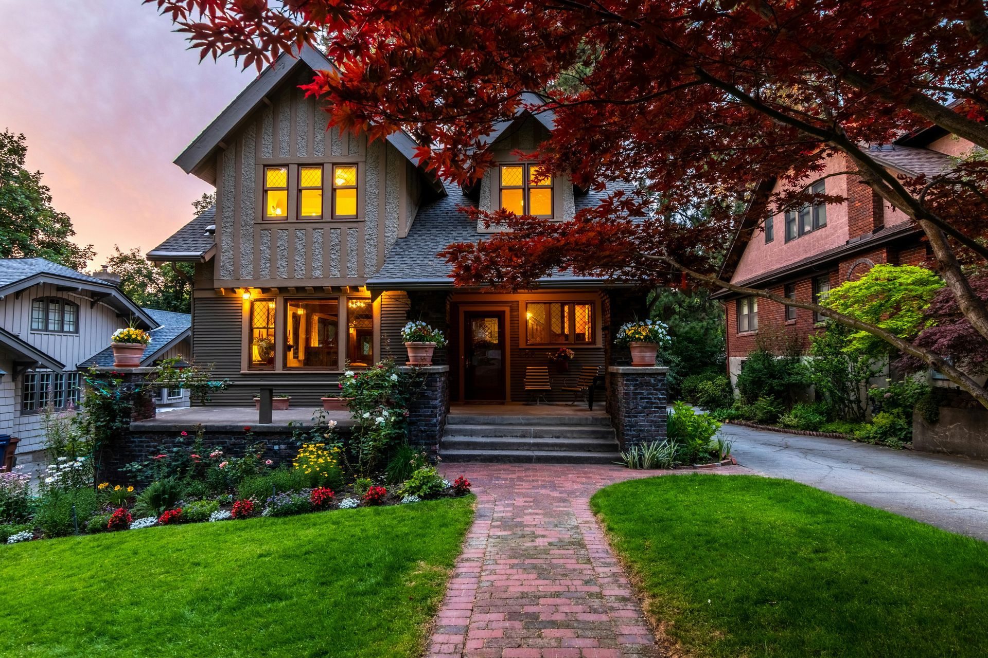A multi-story wooden house with a brick walkway and lawn, illuminated by warm interior lighting at dusk.