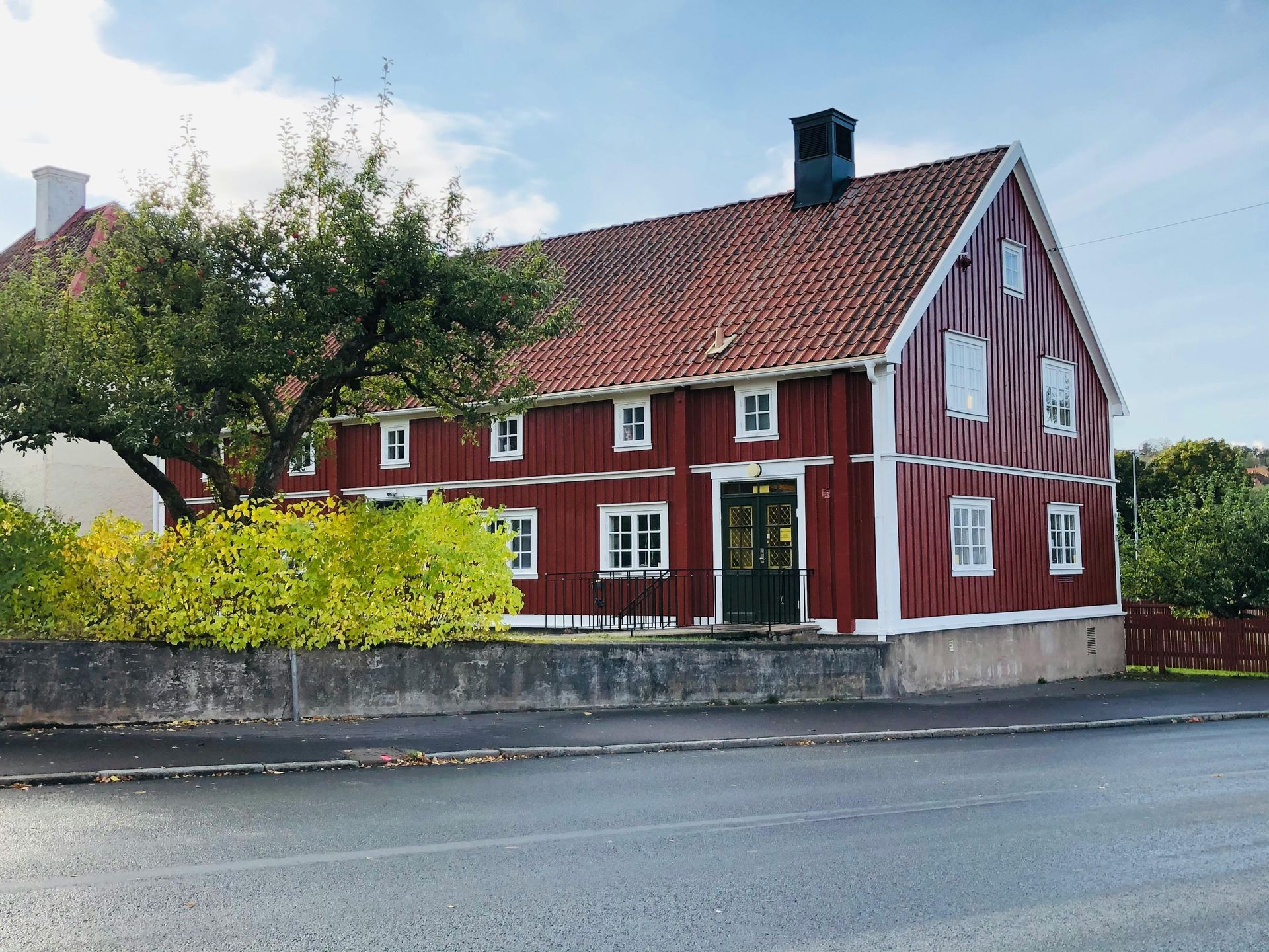 A traditional red Swedish wooden house with white trim, a tiled roof, and a low stone wall along a paved road.