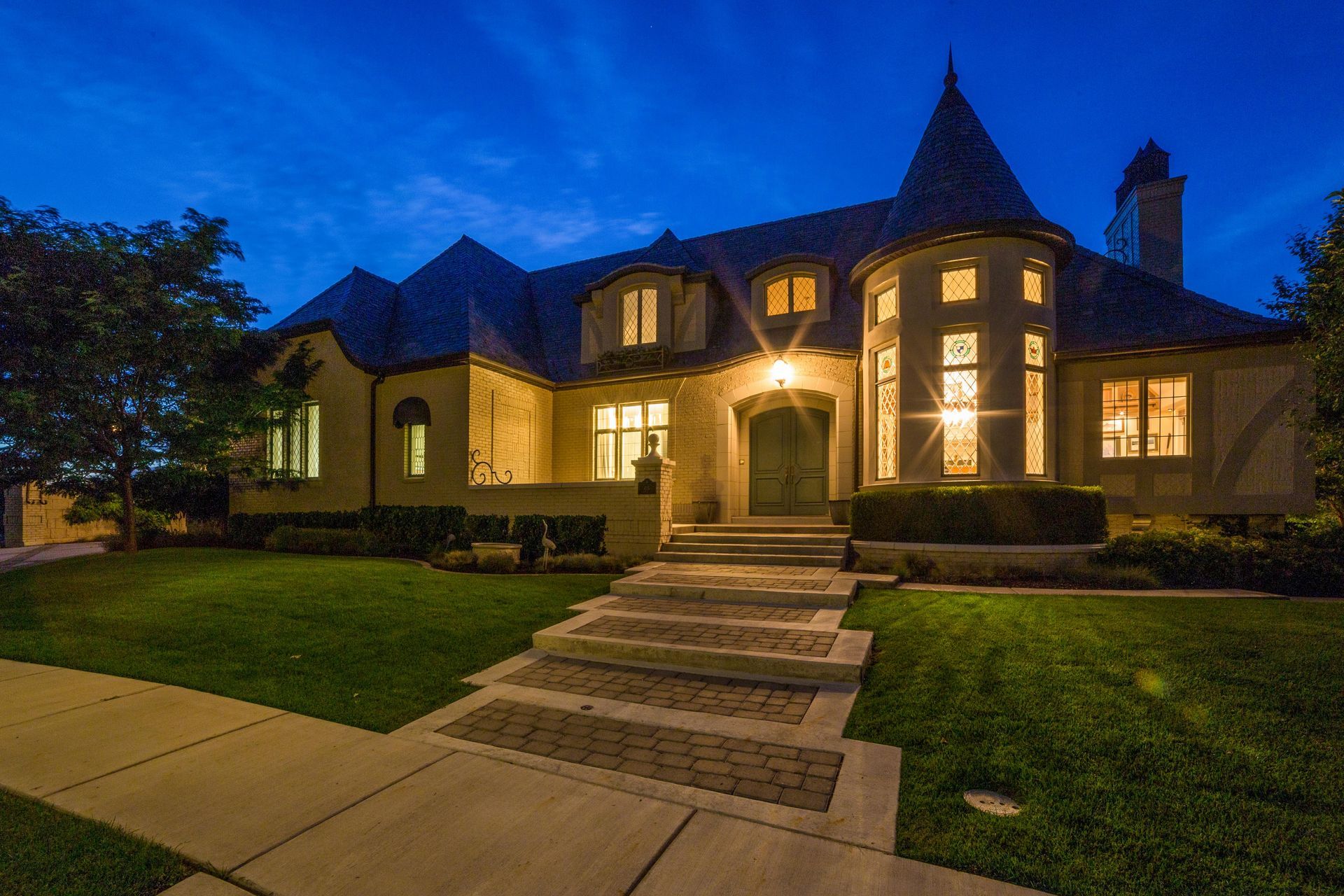 A large, cream-colored house with a turret at dusk, featuring stone steps leading to an illuminated front entrance.