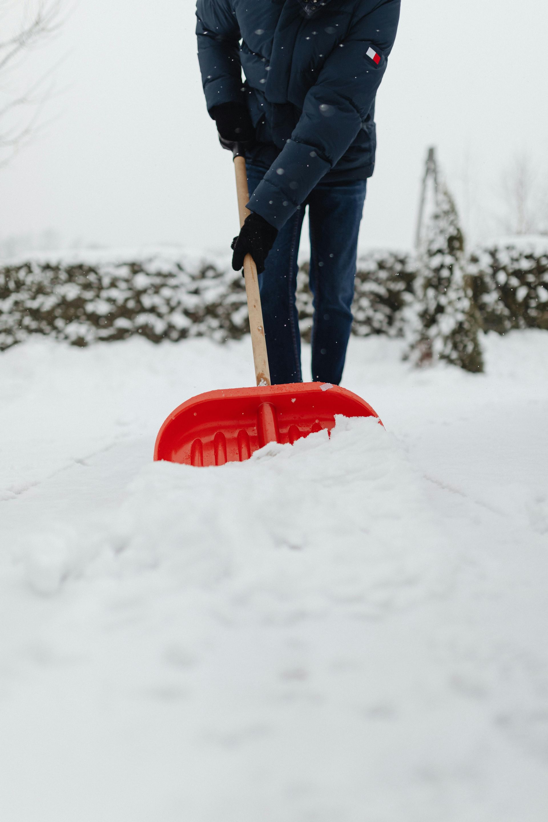 A person in a dark winter coat and gloves uses a bright red shovel to clear a path through deep snow.
