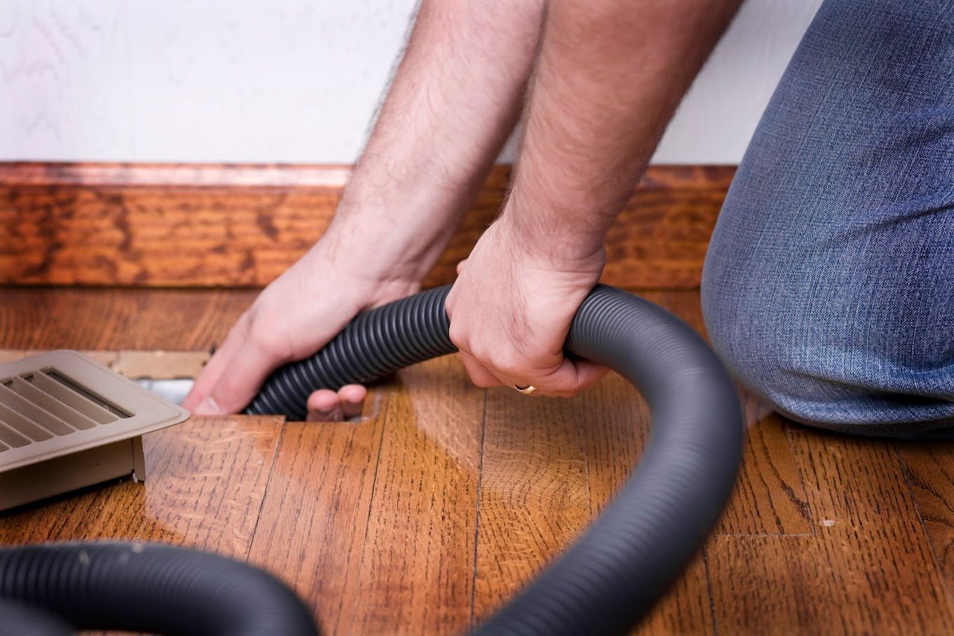 A man is using a vacuum hose to clean a vent on the floor.