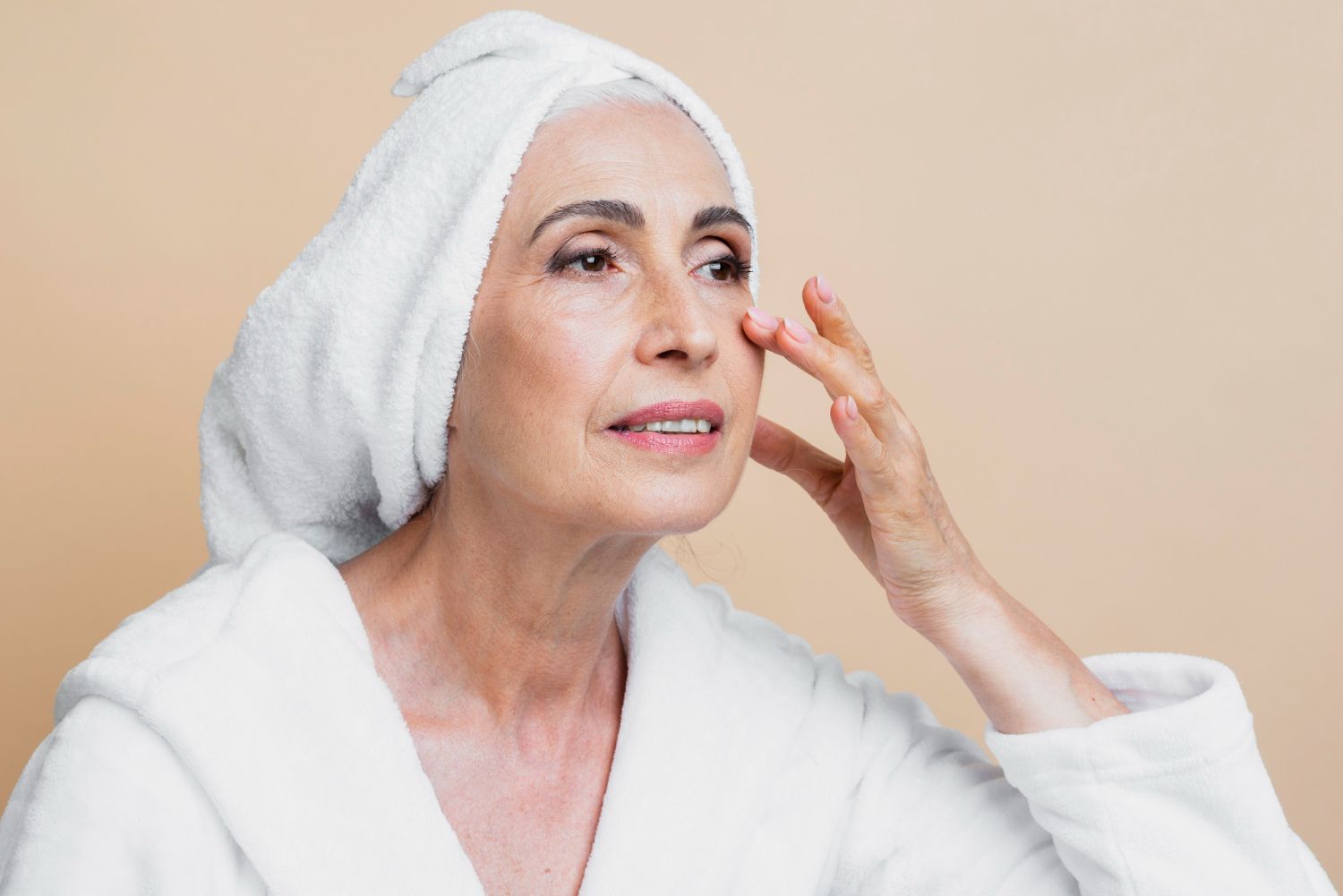 An elderly woman with a towel wrapped around her head is applying lotion to her face.