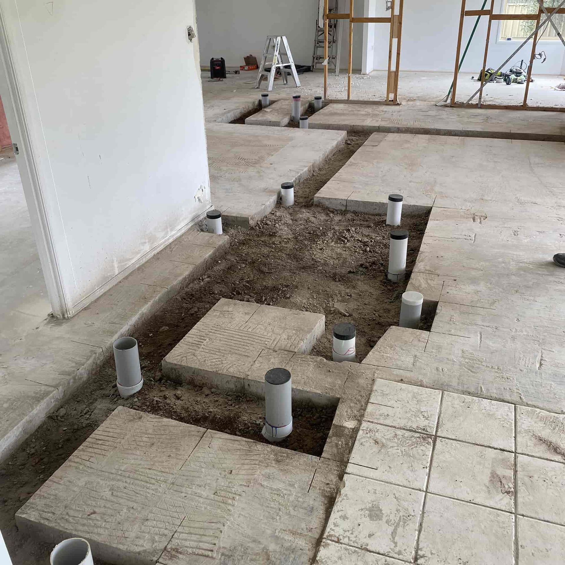 Construction site: concrete floor with trenches, plumbing pipes, concrete blocks, and framing visible.