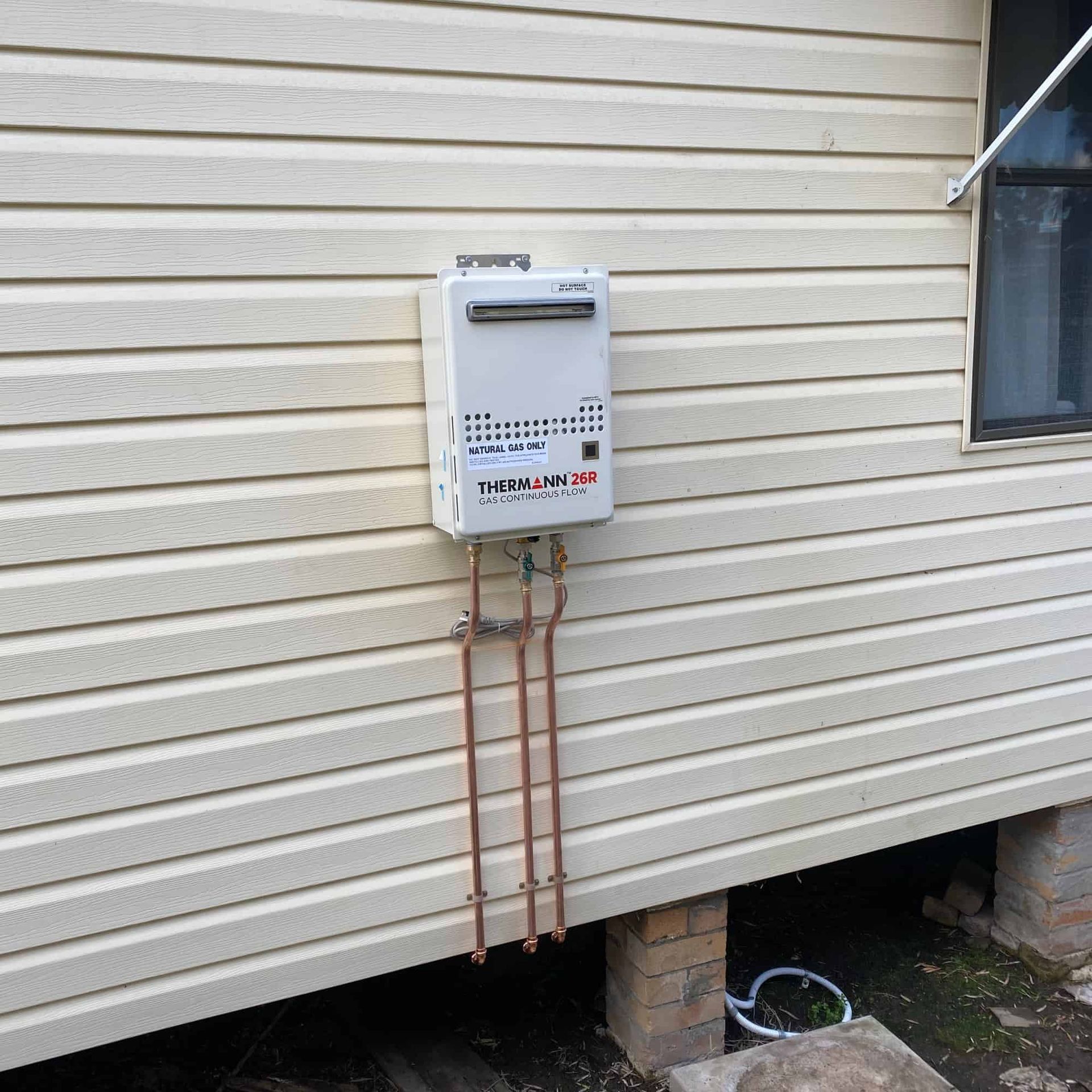 White tank water heater mounted on beige siding with copper pipes.