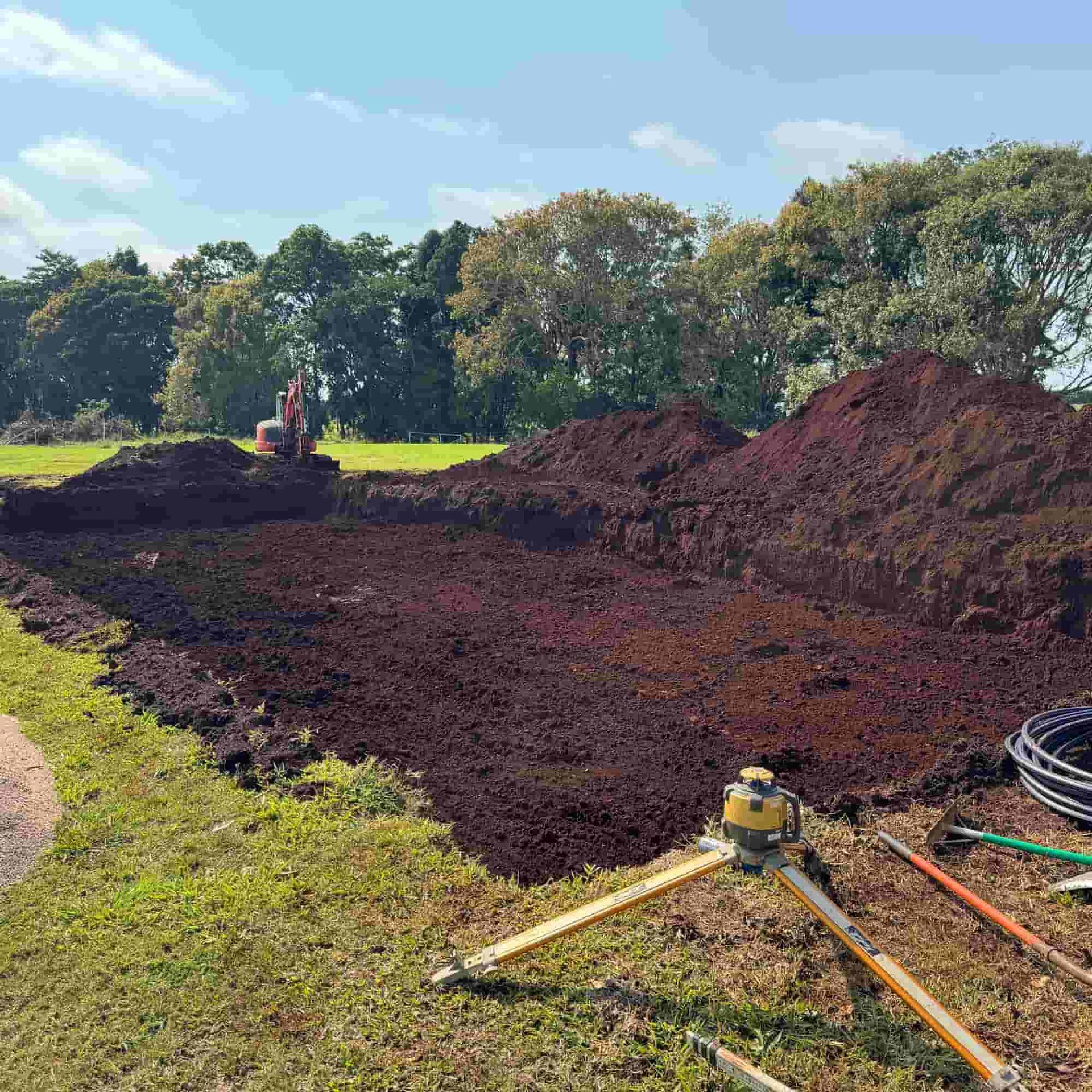 Construction site with dark soil, trees in background, and surveying equipment in the foreground.