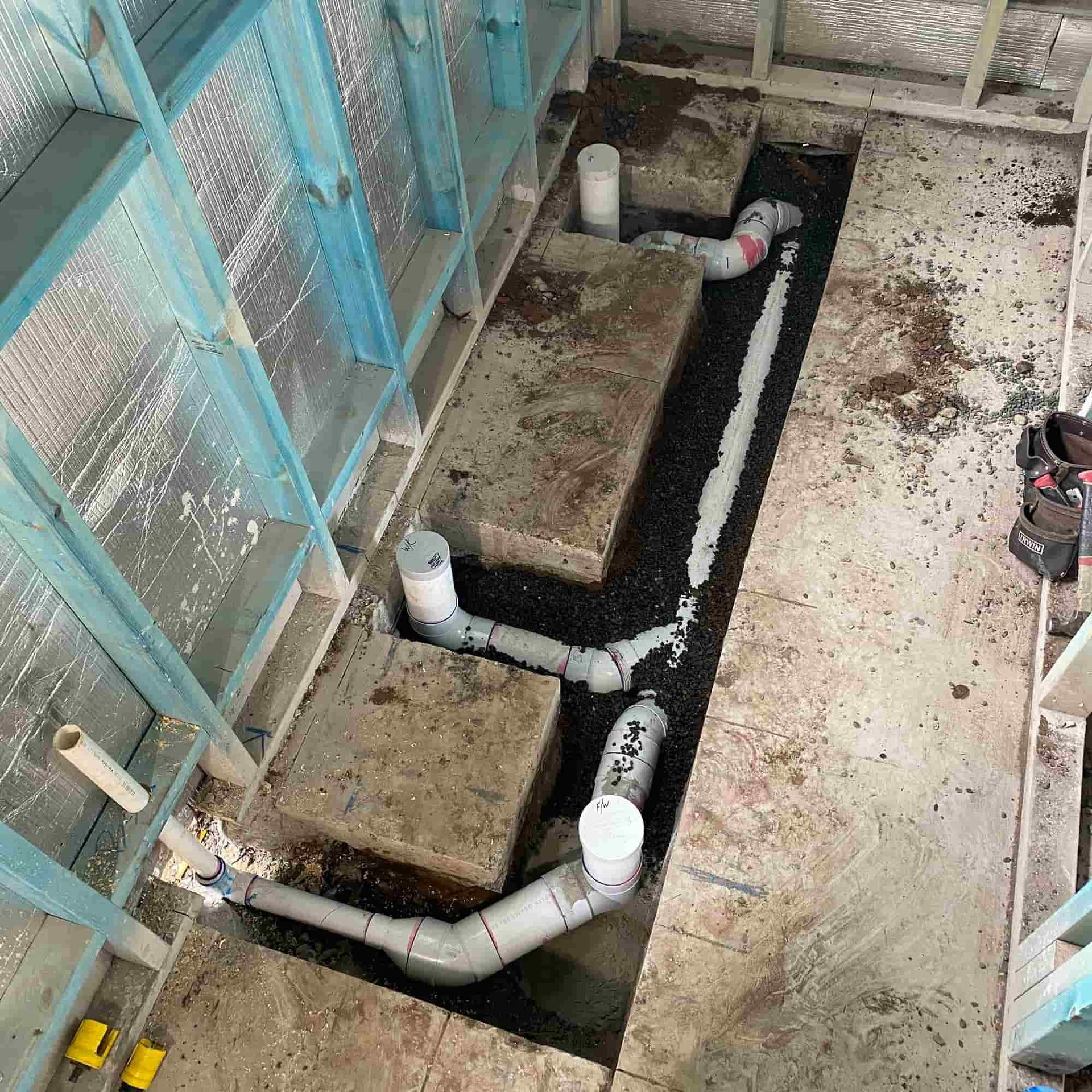 Construction site with exposed plumbing pipes in a trench. Blue framing, insulation, and concrete are visible.