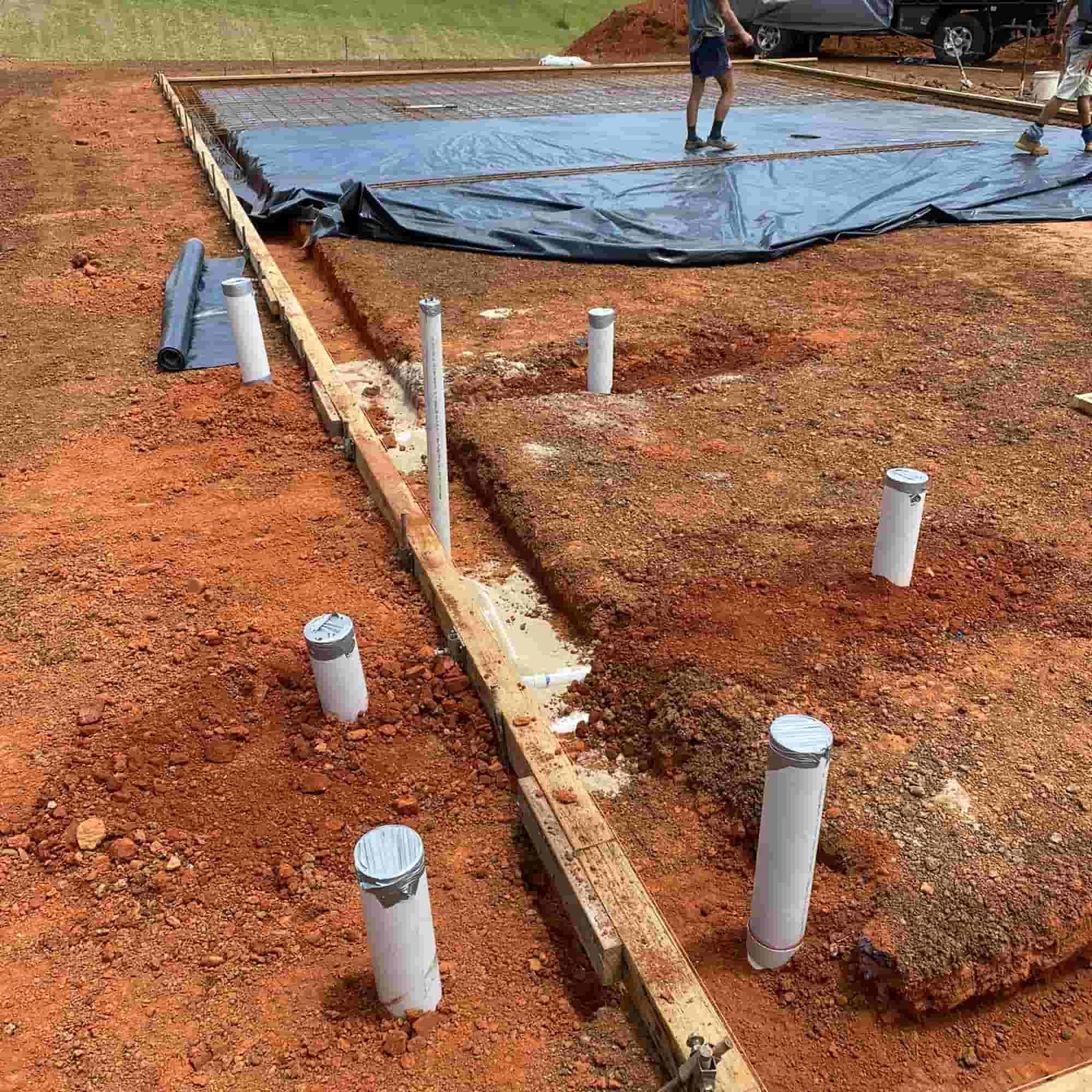 Construction site, ground-level view: foundation prepared with pipes, wood forms, and tarp covering; workers in background.