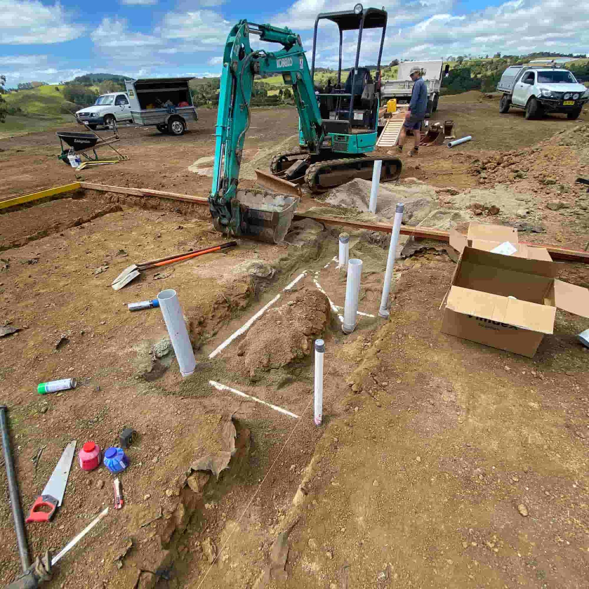 Construction site with a mini-excavator, pipes, and utility vehicles. Person working nearby.