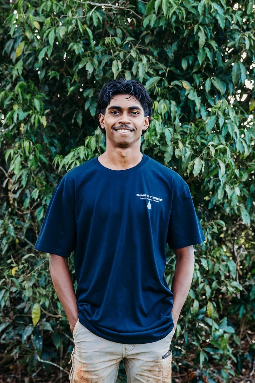 Man in blue shirt and tan pants with hands in pockets smiles in front of a green bush.