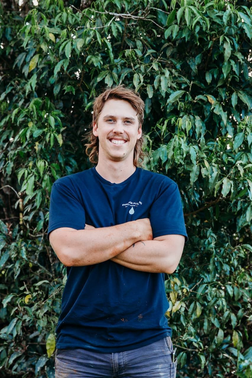 Man smiles, arms crossed, in front of leafy green foliage. He wears a navy shirt and gray pants.