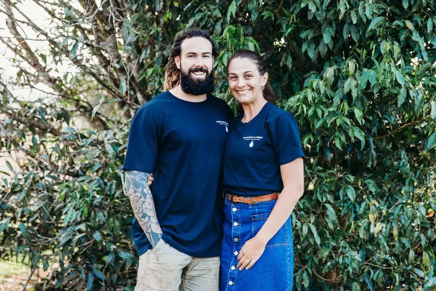 Man with beard and woman in matching blue shirts smile in front of greenery.